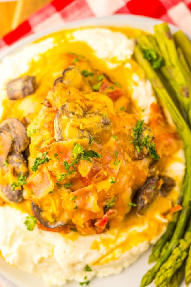 A plate of chicken breast topped with mushrooms and yellow sauce, served over mashed potatoes with a side of steamed asparagus.