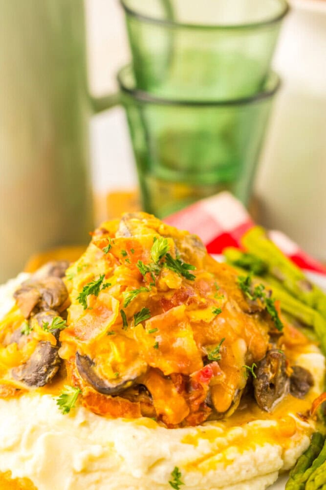 A plate of mashed potatoes topped with a creamy mushroom and vegetable sauce, garnished with parsley, with asparagus spears and stacked green glasses in the background.