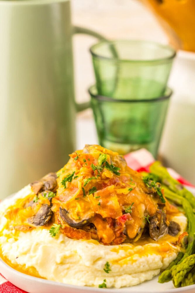 A plate of mashed potatoes topped with a creamy mushroom and vegetable sauce, garnished with parsley, served with a side of asparagus. Green glasses and a mug are in the background.