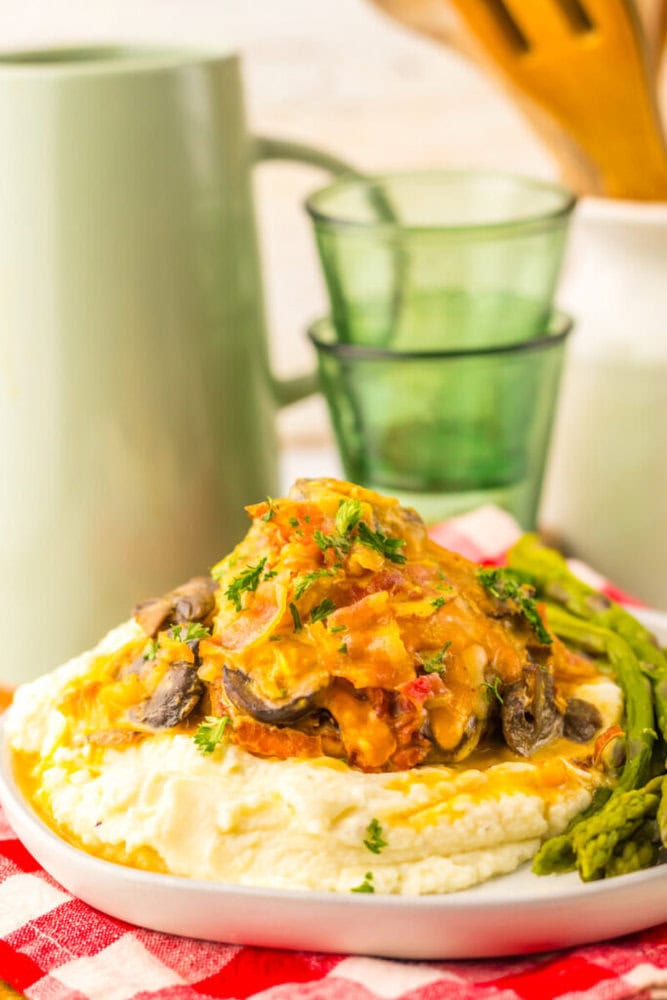 A serving of mashed potatoes topped with a creamy chicken and vegetable mixture, garnished with herbs, and a side of asparagus on a white plate.