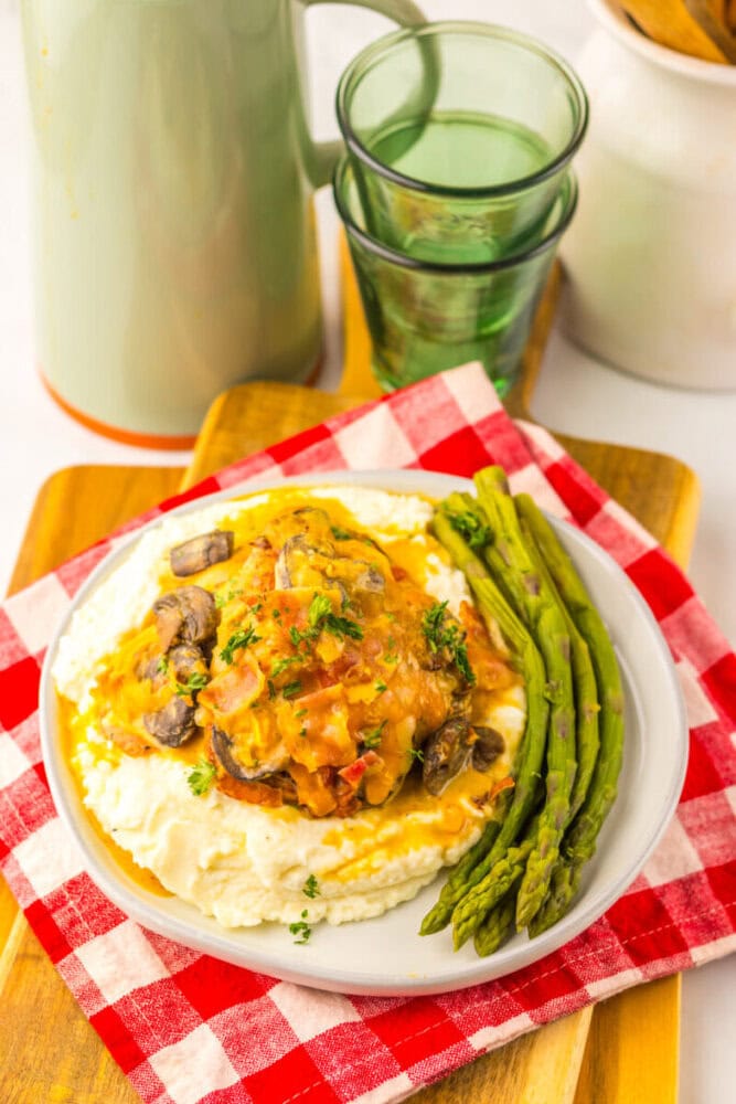 A plate of creamy mashed potatoes topped with a mushroom and vegetable sauce, served with steamed asparagus, placed on a red checkered napkin beside two stacked green glasses.