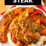 A plate of Slow Cooker Swiss Steak with rich gravy, carrots, and onions served over mashed potatoes on a red checked cloth.