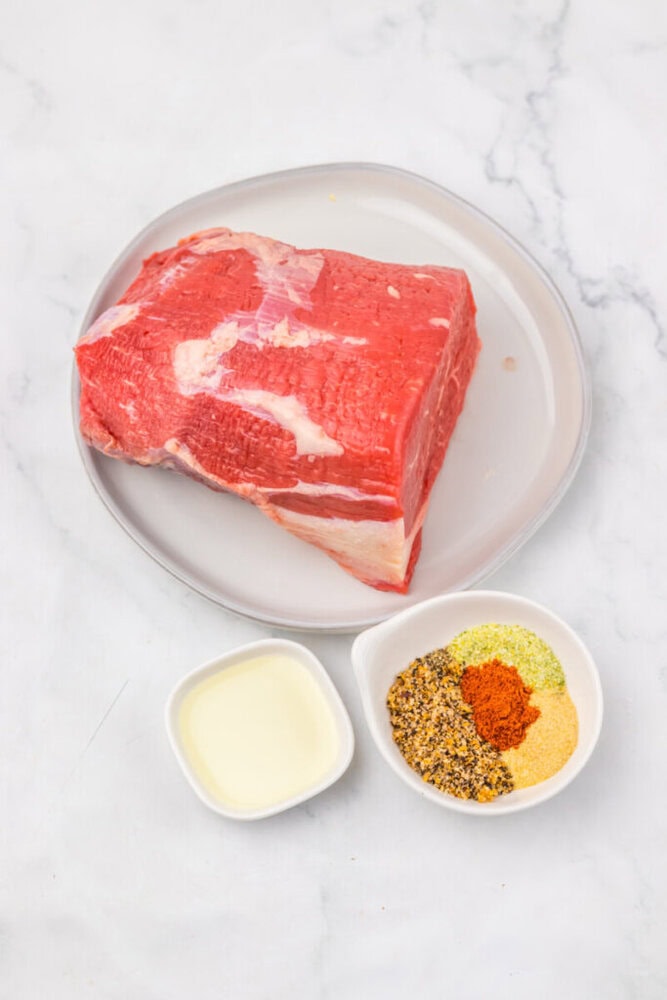 A raw beef roast on a plate, next to a small bowl of oil and a small bowl with assorted dry seasonings on a white surface.
