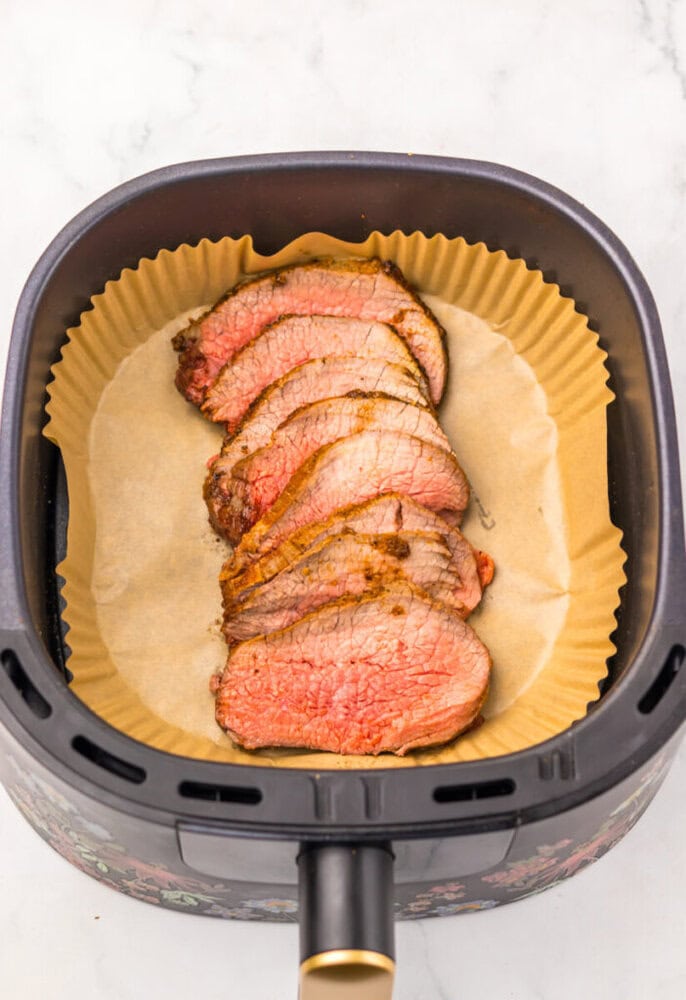Sliced cooked roast beef arranged on parchment paper inside an air fryer basket.