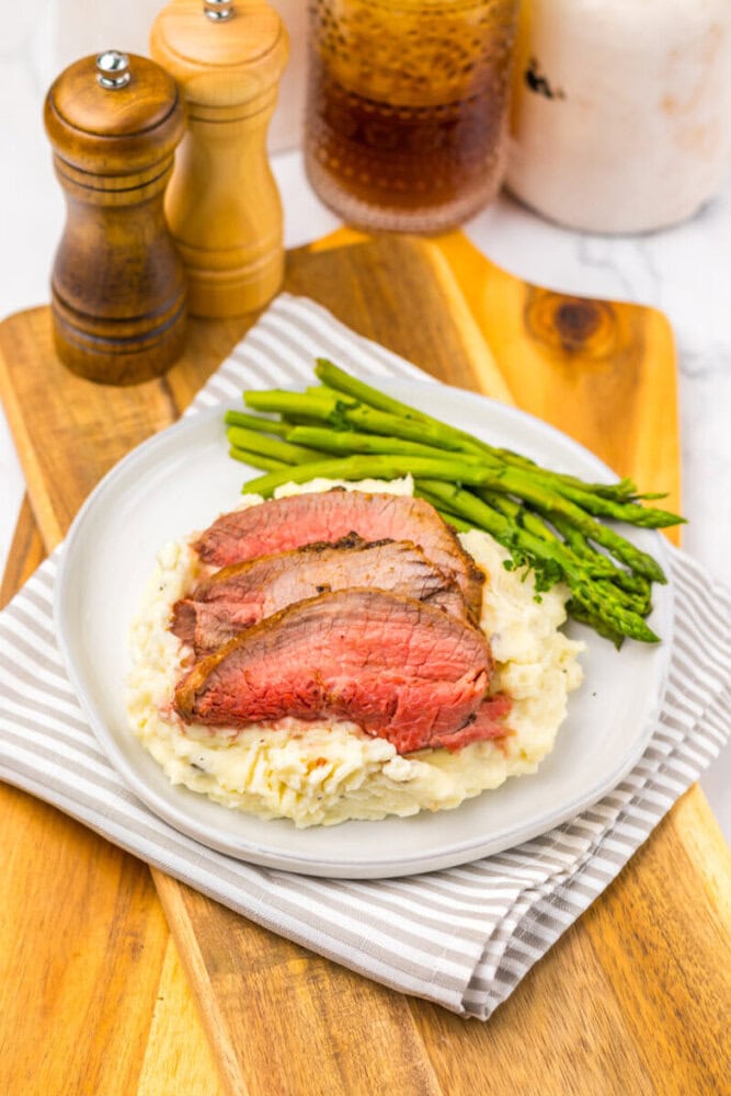 A plate with sliced steak on mashed potatoes and asparagus, placed on a striped napkin atop a wooden board. Salt and pepper shakers and a drink are in the background.