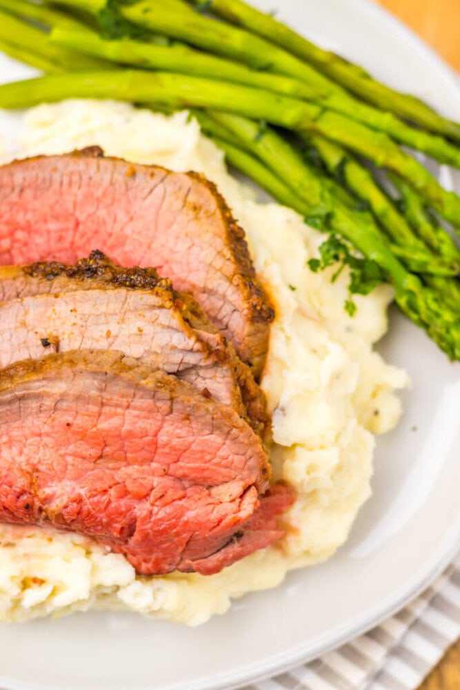 Three slices of medium-rare roast beef on mashed potatoes with a side of asparagus on a white plate.