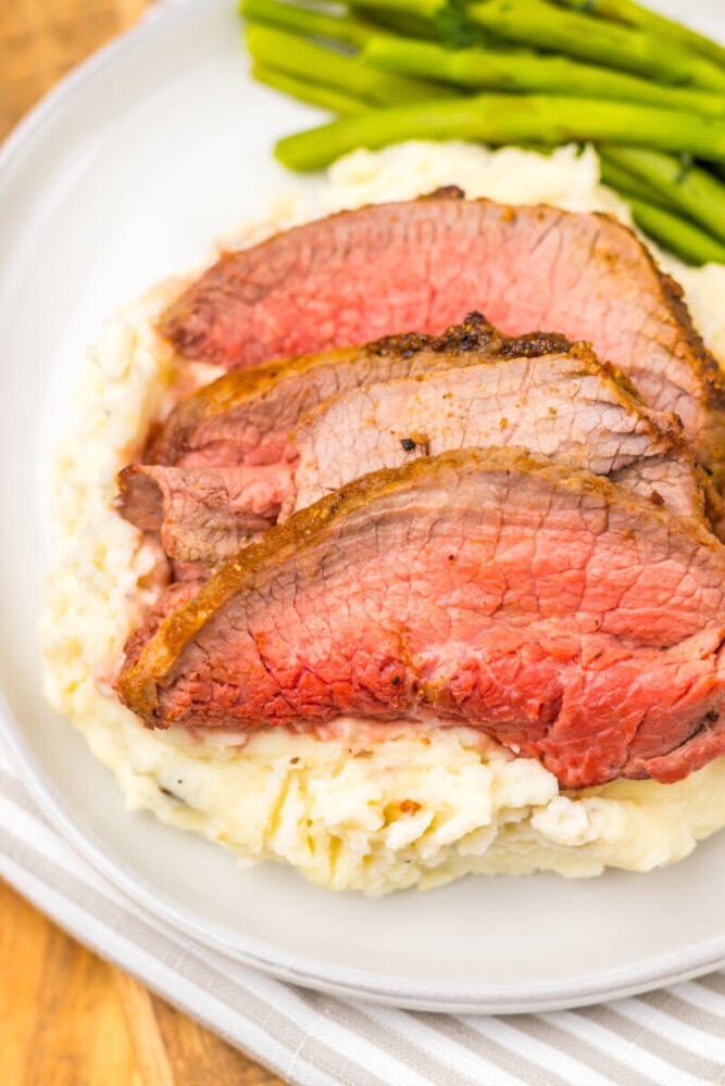 Three slices of medium-rare roast beef served on mashed potatoes with a side of green beans on a white plate.