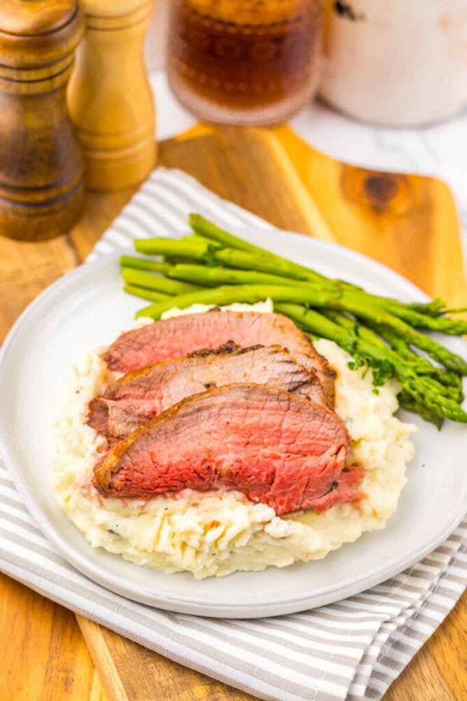 Three slices of medium-rare steak served on mashed potatoes with a side of asparagus on a white plate, placed on a striped napkin and wooden board.