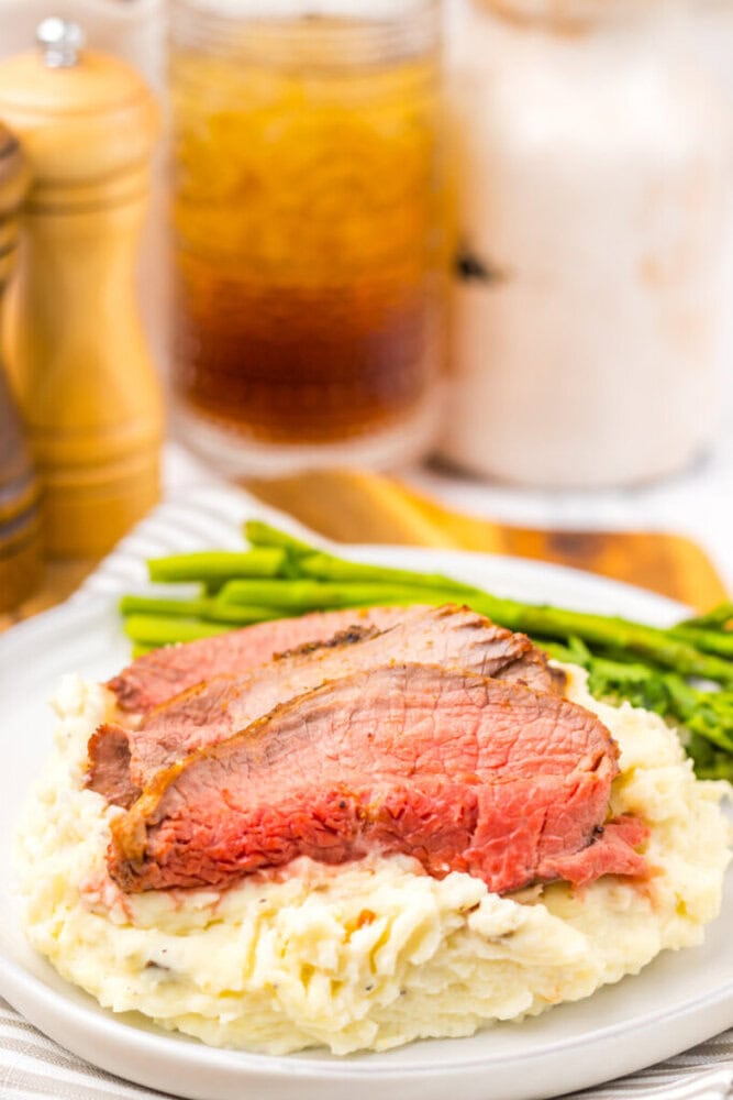 Sliced roast beef served on mashed potatoes with a side of asparagus, with drinks and condiments in the background.