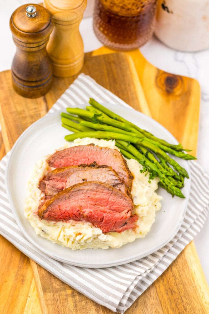 Sliced roast beef on mashed potatoes with steamed asparagus, served on a white plate with a striped napkin, on a wooden board.