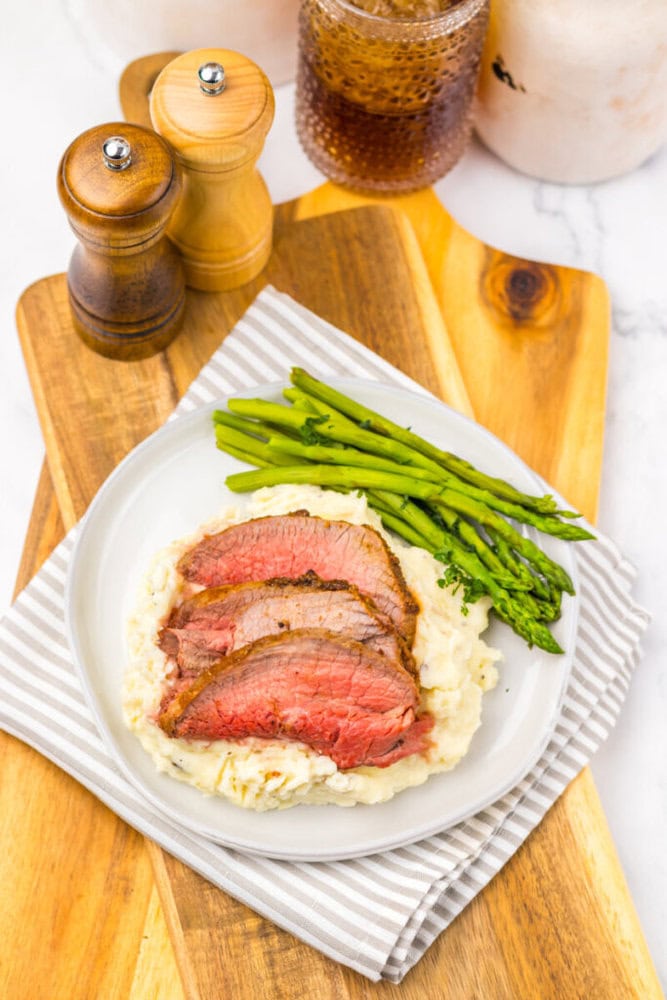 A white plate with three slices of roast beef on mashed potatoes and asparagus spears, set on a striped napkin with salt and pepper shakers and a drink in the background.