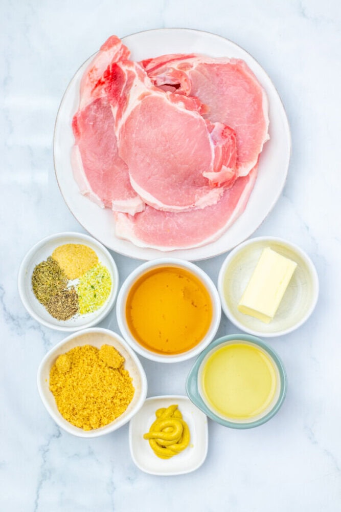 Raw pork chops on a plate with small bowls containing mustard, butter, broth, honey, seasonings, and a yellow powder, arranged on a white surface.