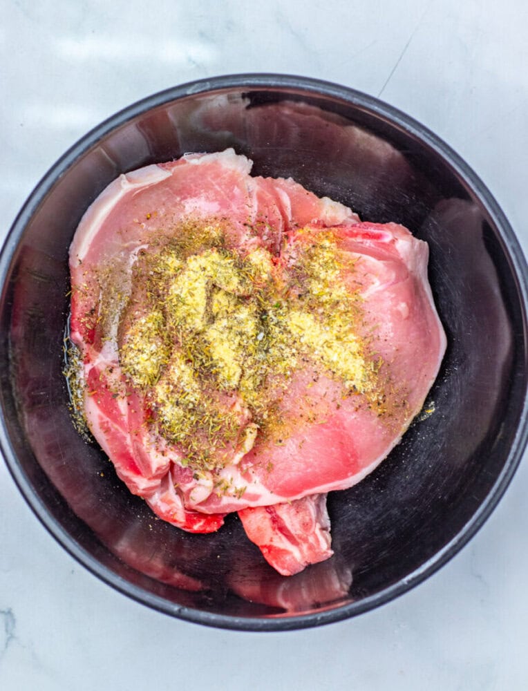 Raw pork chops in a black bowl, topped with a mixture of dried herbs and seasonings, on a light-colored surface.