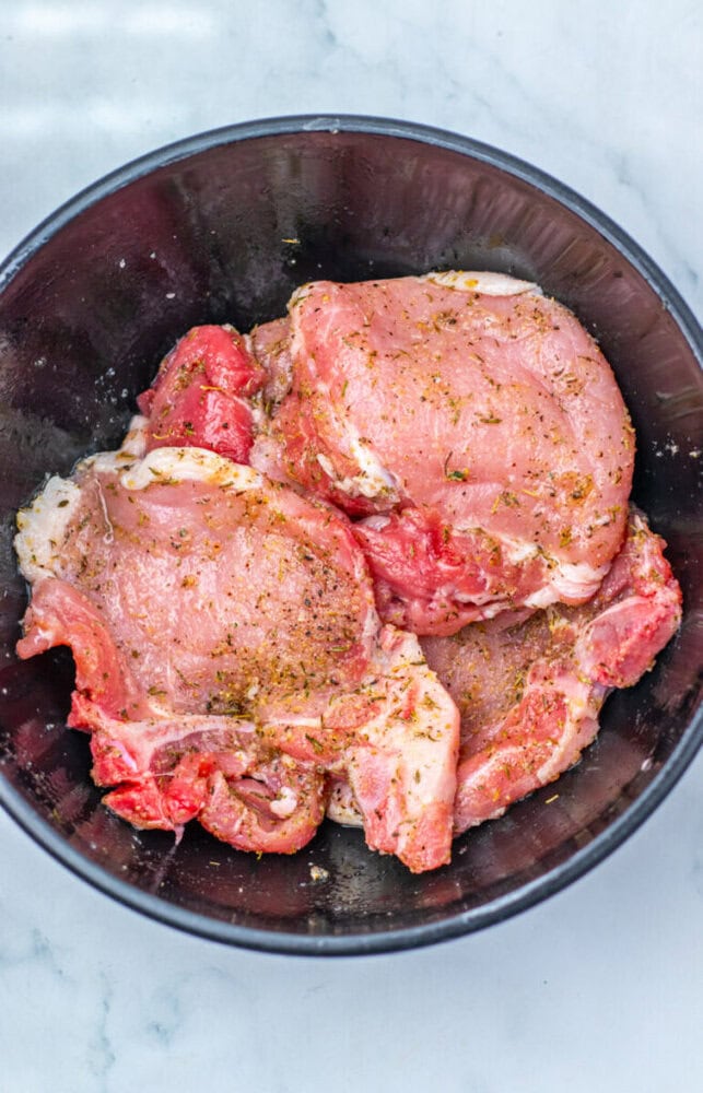 Raw, seasoned pork steaks placed in a black bowl on a light-colored surface.