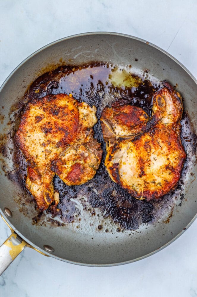 Two cooked, seasoned pork chops in a frying pan with browned bits and oil on a light surface.