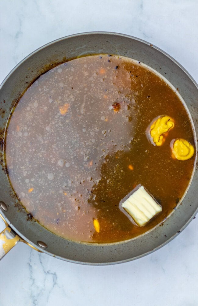 A frying pan with brown liquid, a pat of butter, and two dollops of yellow mustard on a light marble surface.
