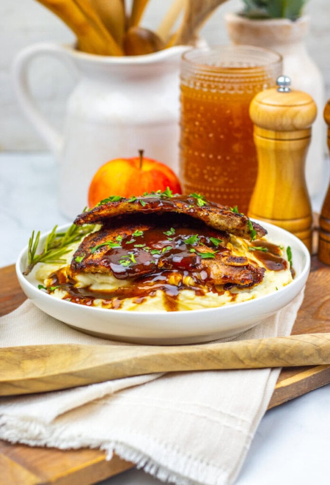 A plate of mashed potatoes topped with two pieces of grilled meat, brown sauce, and herbs, with an apple, a glass of drink, and condiments in the background.