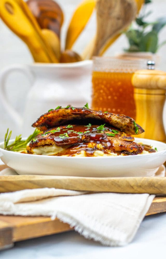 A plate with two stacked grilled sandwiches topped with sauce and herbs, set on a white napkin with kitchen utensils and a pepper mill in the background.