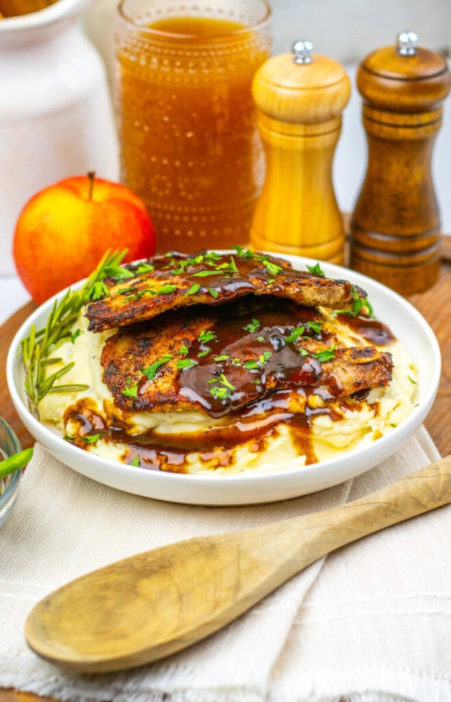 A plate of mashed potatoes topped with grilled pork chops and brown gravy, garnished with herbs, next to a wooden spoon, salt, pepper, an apple, and a jar of sauce.