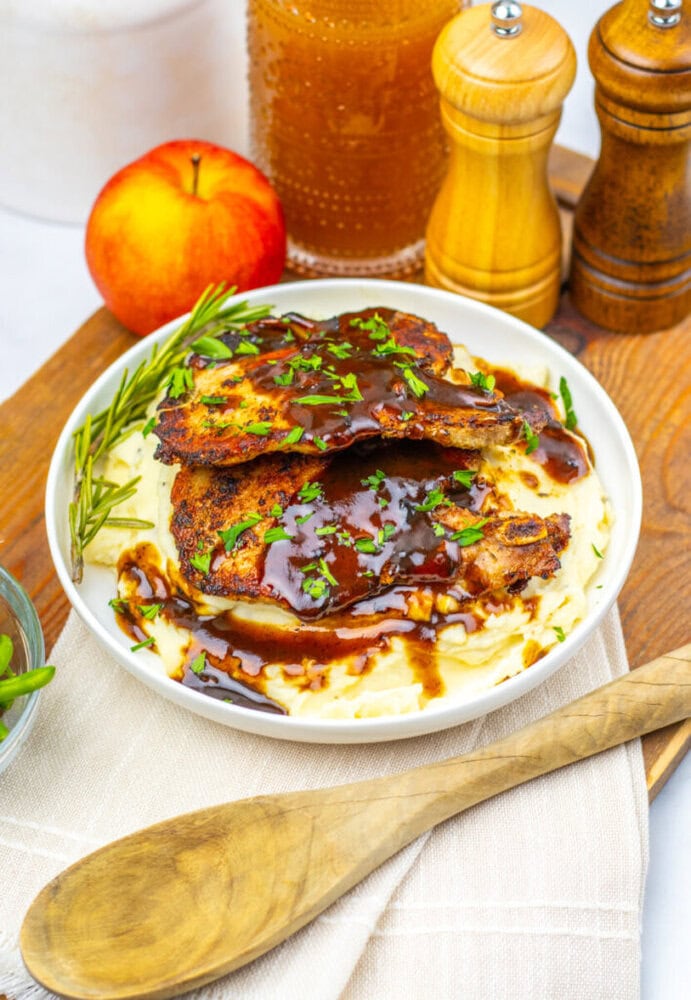 A plate of mashed potatoes topped with grilled chicken and brown sauce, garnished with herbs, sits on a wooden board with a peach, honey jar, salt and pepper shakers, and a wooden spoon.