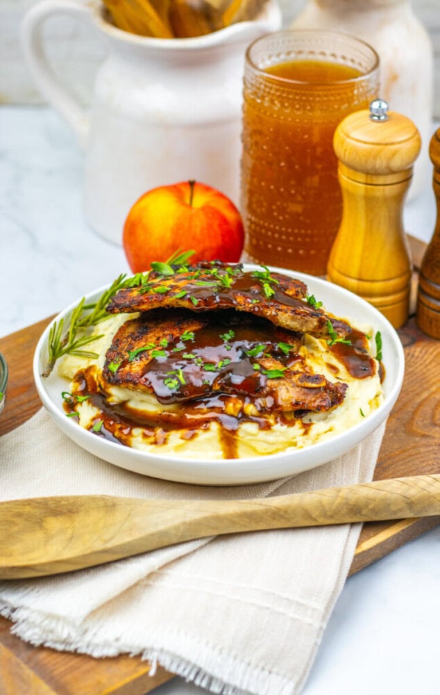 A plate of grilled pork chops with brown gravy on mashed potatoes, garnished with herbs, with a peach, pepper grinder, and a drink in the background.
