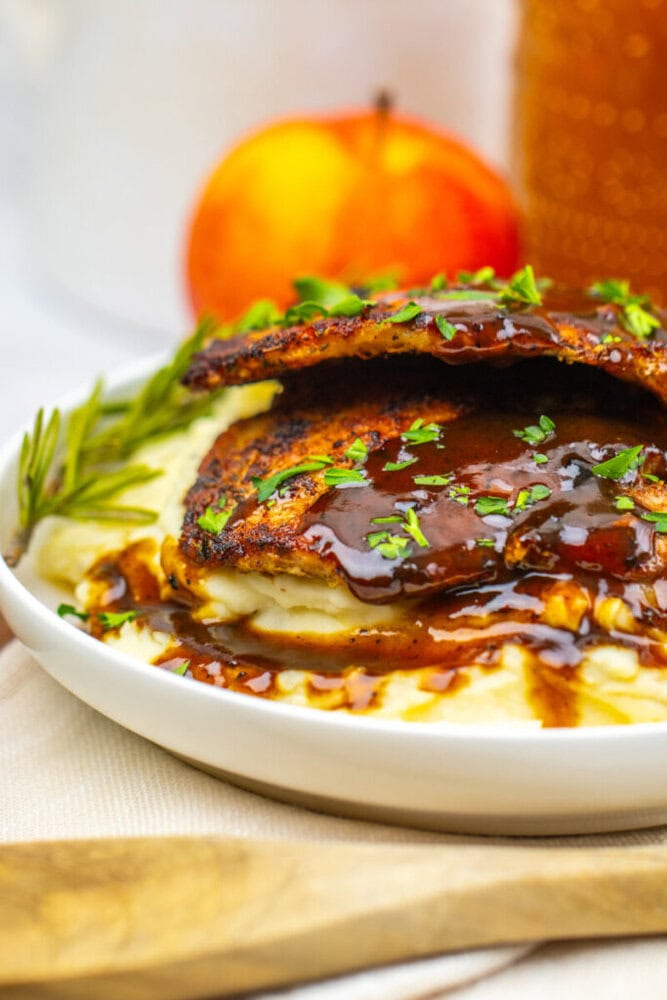 A plate of mashed potatoes topped with grilled chicken breasts, brown sauce, chopped herbs, and a sprig of rosemary, with an apple and honey jar in the background.