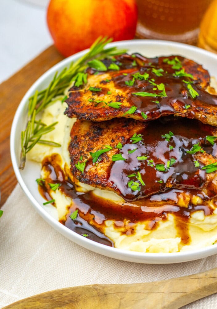 Grilled chicken breasts with brown sauce served over mashed potatoes, garnished with chopped herbs and a sprig of rosemary, with a peach and drink in the background.