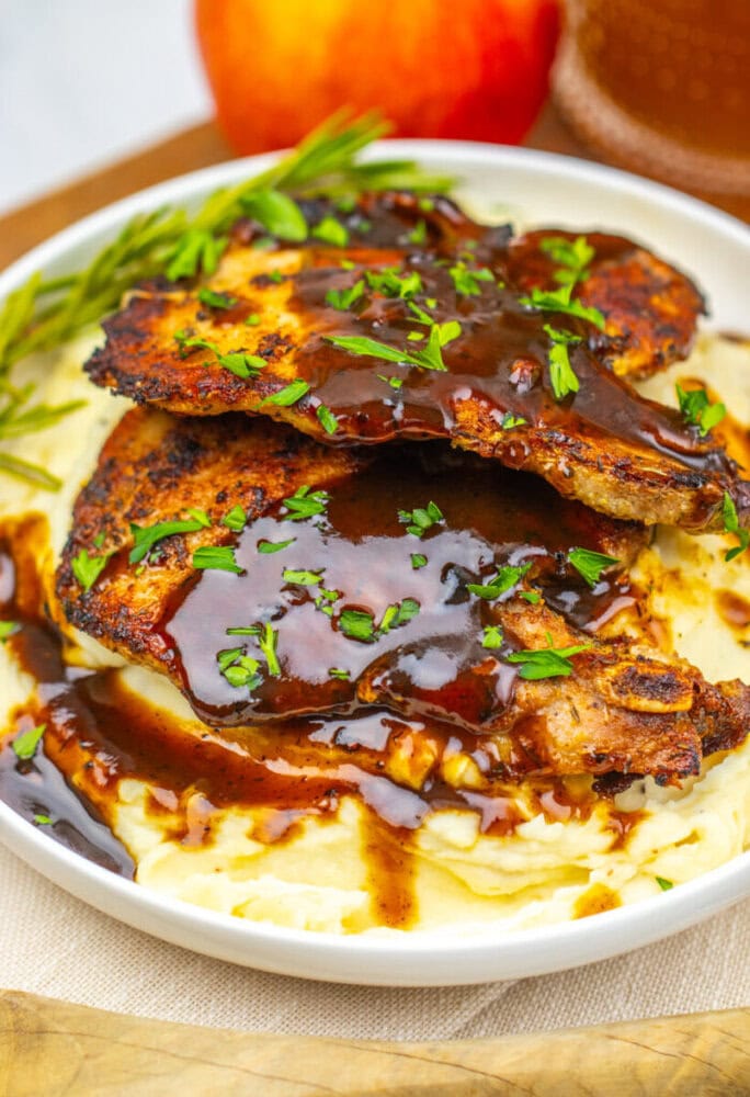 Grilled pork chops with dark sauce on mashed potatoes, garnished with chopped herbs and a sprig of rosemary, served in a white bowl.