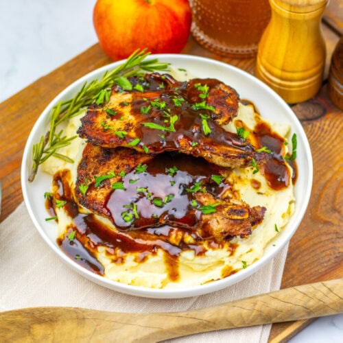 A plate of mashed potatoes topped with two pieces of grilled meat and brown gravy, garnished with herbs, with green beans, an apple, and seasonings nearby.