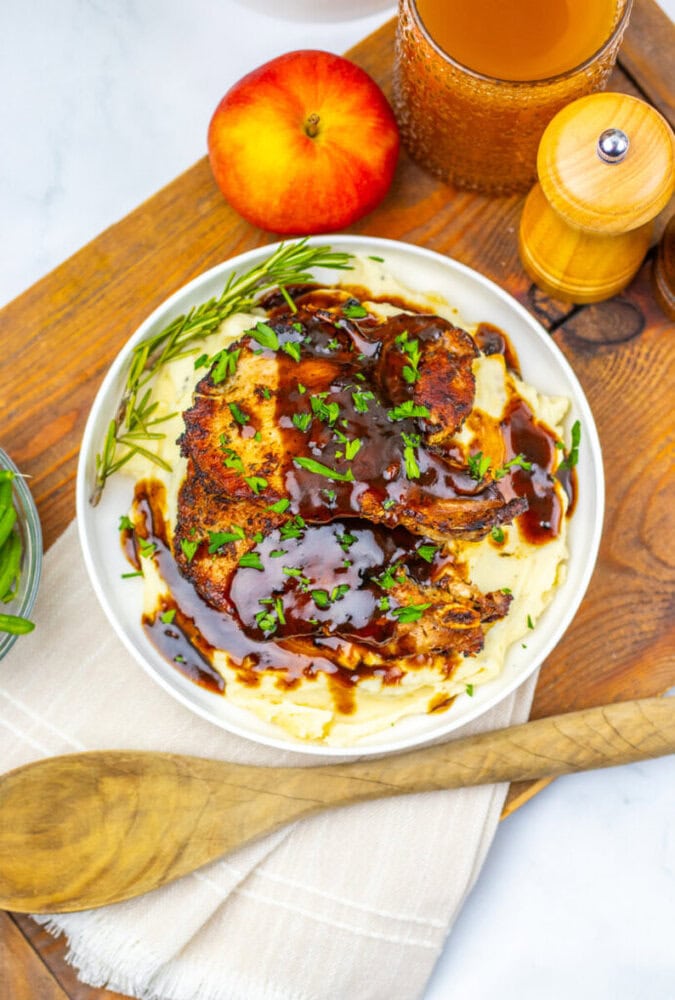 A bowl of mashed potatoes topped with grilled chicken, brown sauce, and chopped herbs, placed on a wooden board with a peach, glass of juice, pepper grinder, and a wooden spoon nearby.