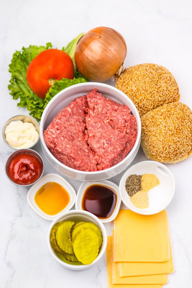 Ingredients for burgers arranged on a white surface, including ground beef, cheese slices, burger buns, pickles, tomato, lettuce, onion, sauces, and seasonings.