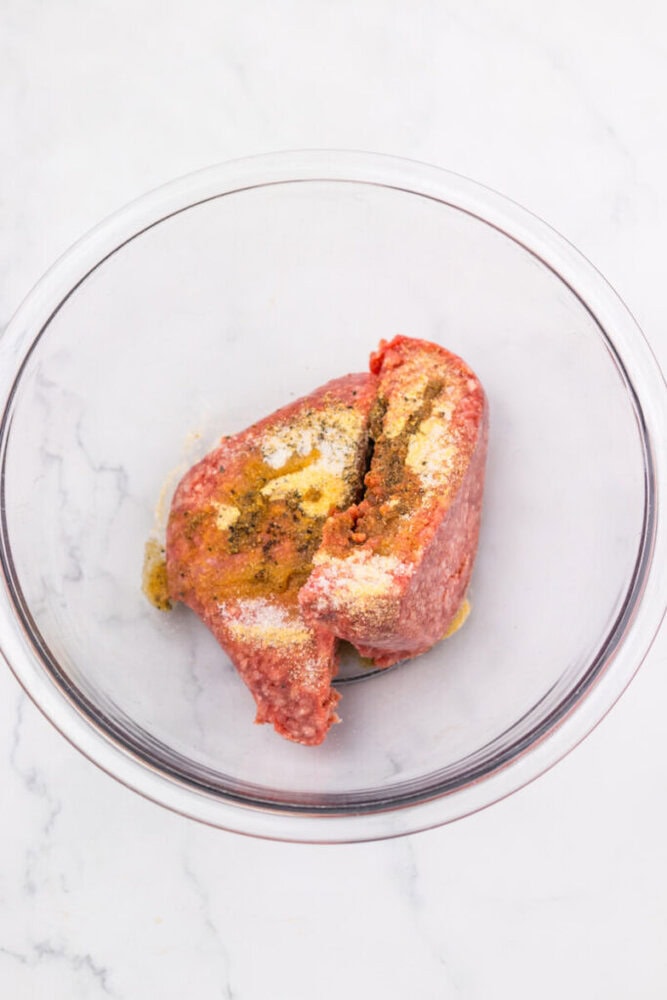 Raw ground beef with various spices sprinkled on top in a clear glass mixing bowl on a white marble surface.