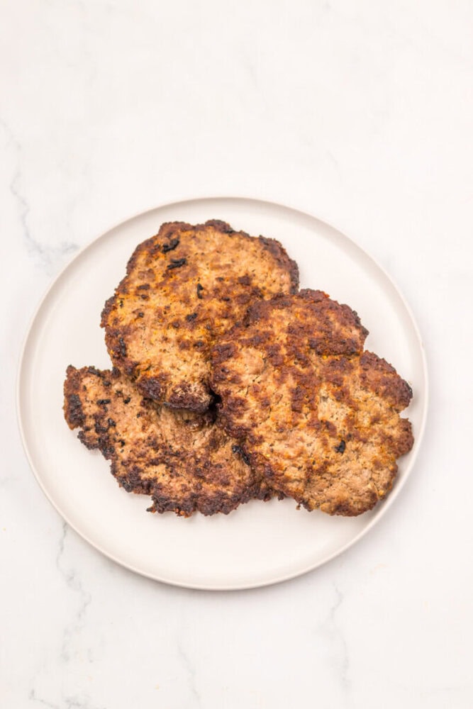 Three cooked ground beef patties are arranged on a white plate, set on a light marble surface.