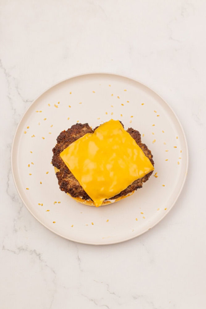 A plate with a cooked hamburger patty topped with a slice of melted cheddar cheese, with sesame seeds scattered around on the plate.