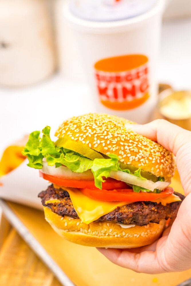 A hand holds a Burger King cheeseburger with lettuce, tomato, onion, and pickles on a sesame seed bun; Burger King cup and fries in the background.