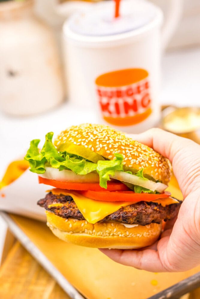 A hand holding a cheeseburger with lettuce, tomato, onion, and cheese in front of a Burger King cup.