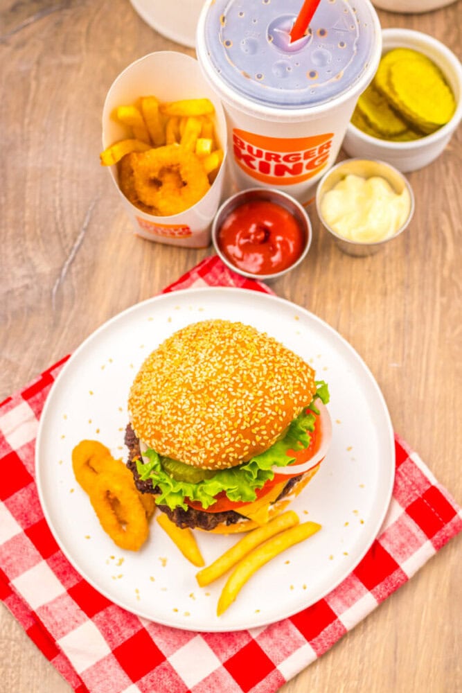 A sesame seed burger with lettuce, tomato, and cheese, served with fries, onion rings, dipping sauces, pickles, and a soda on a red checkered napkin.