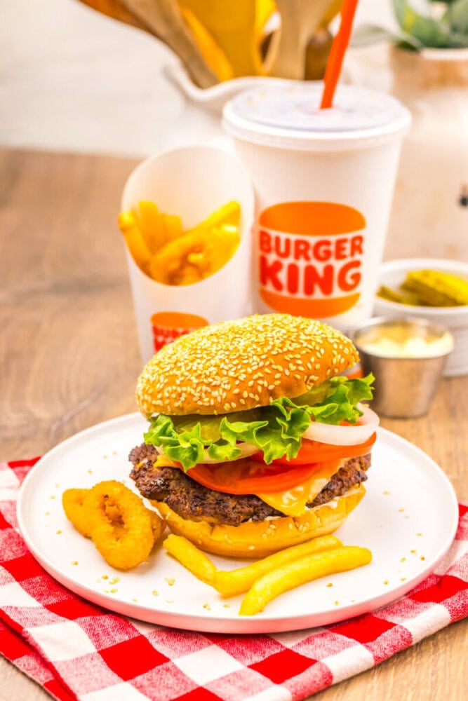 A Burger King meal with a cheeseburger, french fries, onion rings, and a soft drink on a white plate, served on a red checkered napkin.