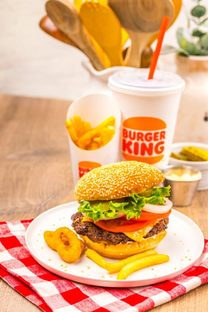 A Burger King meal with a cheeseburger, French fries, onion rings, a soft drink, and condiments on a white plate over a red and white checkered napkin.