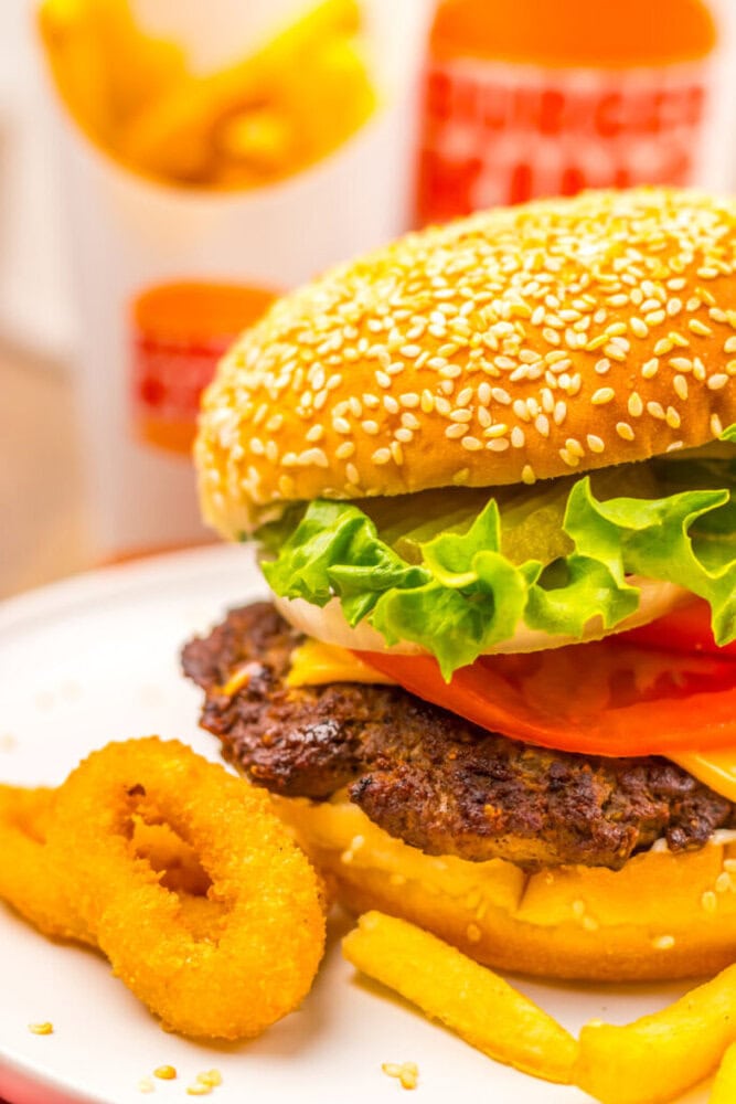 A sesame seed hamburger with lettuce, tomato, cheese, and pickles, served with French fries and an onion ring on a white plate. A cup of fries and a drink are in the background.