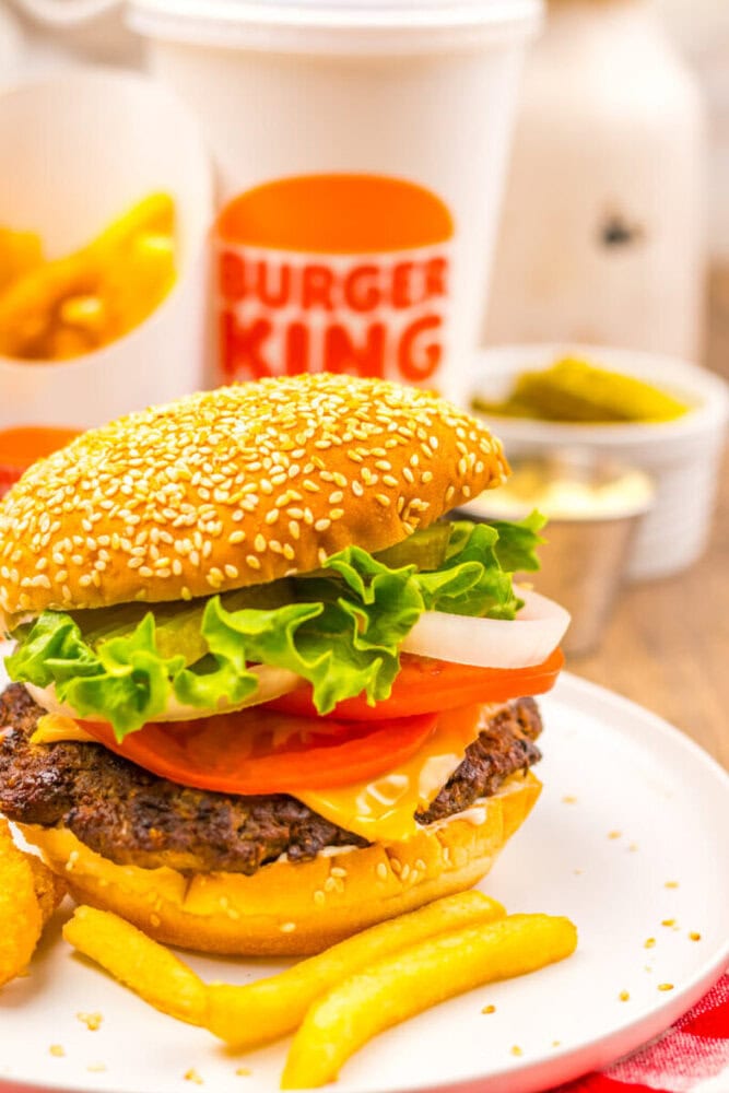A cheeseburger with lettuce, tomato, onions, and cheese on a sesame bun, served with French fries. A Burger King cup and fries container are in the background.