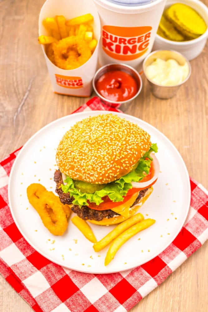 Burger King meal with a cheeseburger, French fries, onion rings, ketchup, mayonnaise, pickles, and a drink on a checkered napkin.