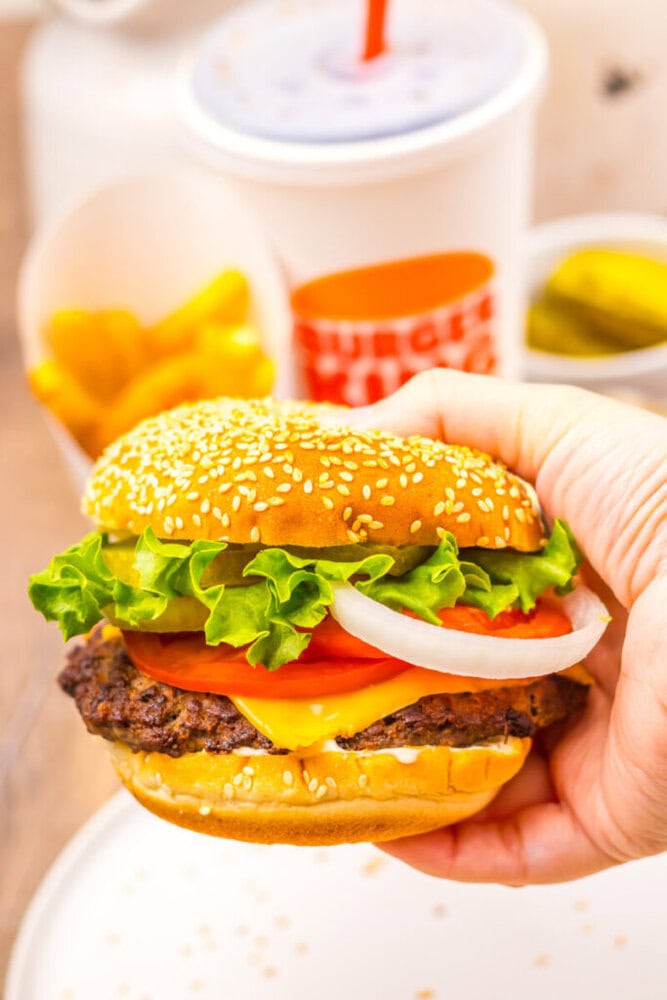 A hand holding a cheeseburger with lettuce, tomato, onion, and cheese in a sesame seed bun; fries, pickles, and a drink are in the background.