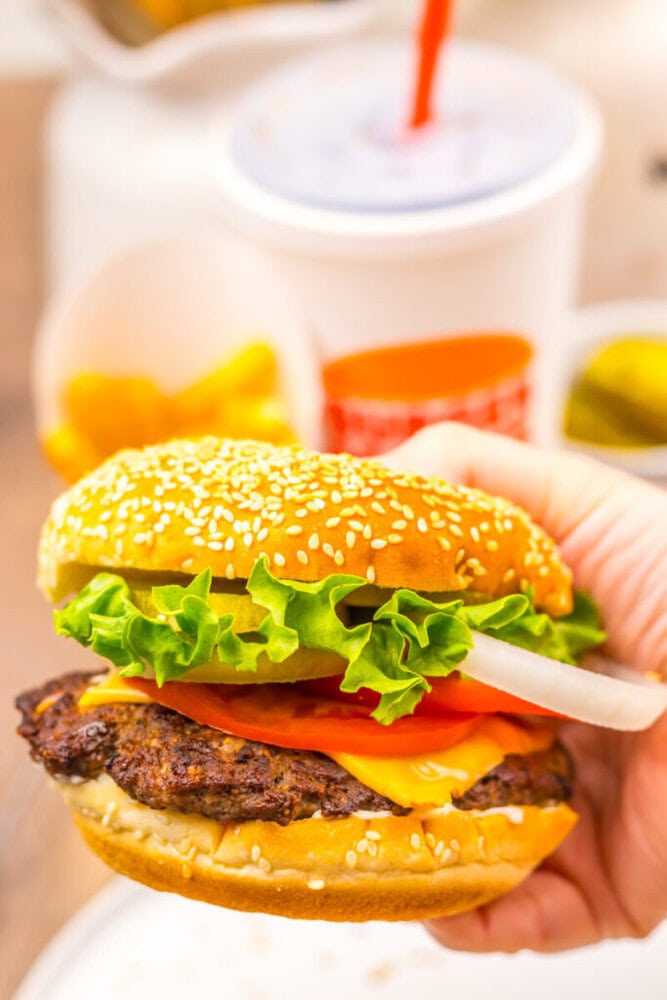 A hand holds a cheeseburger with lettuce, tomato, onion, and cheddar on a sesame bun; a cup with a straw and fries are in the background.