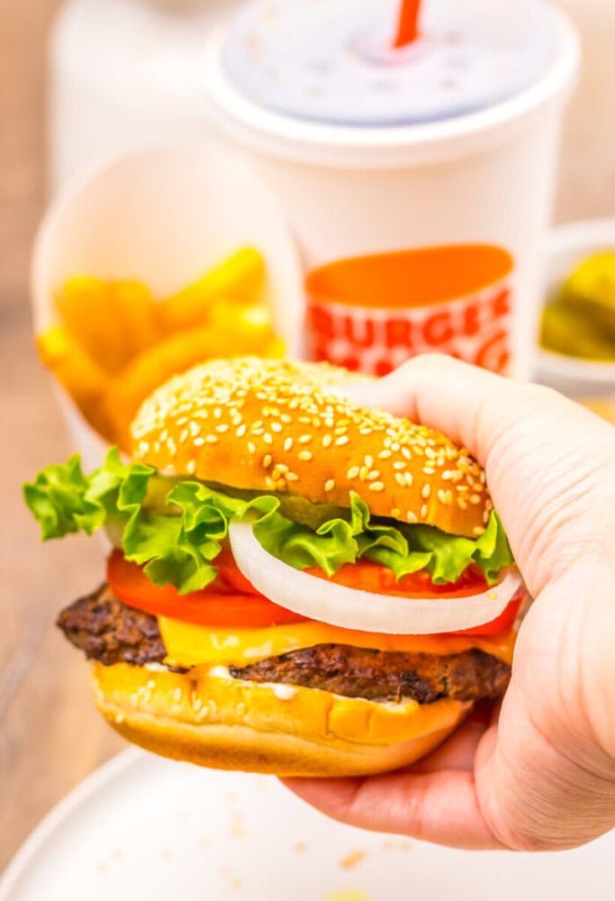 A hand holds a cheeseburger with lettuce, tomato, onion, and cheese. In the background are French fries, a cup with a straw, and a small dish of pickles.