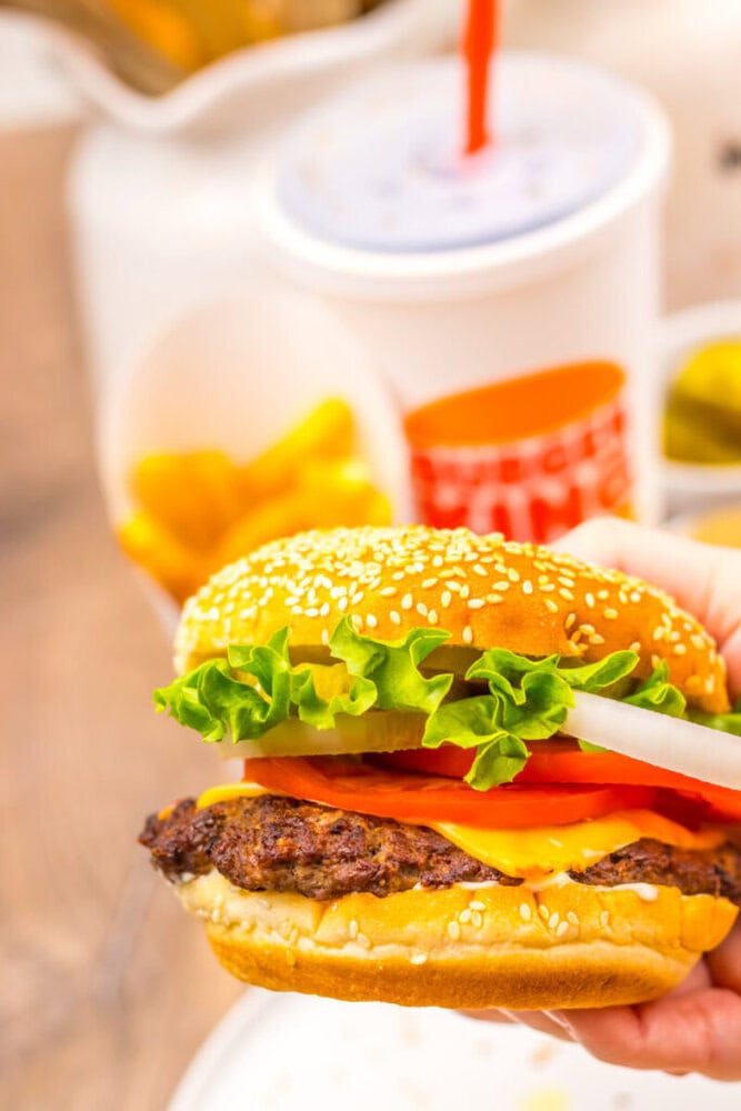A hand holds a cheeseburger with lettuce, tomato, onion, and a beef patty in a sesame seed bun. In the background are fries, pickles, and a soft drink.
