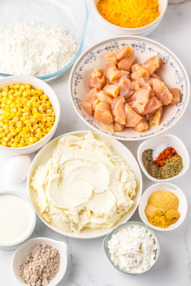 Bowls containing cubed raw chicken, shredded cheese, corn, flour, mashed potatoes, spices, egg, milk, and other baking ingredients arranged on a white surface.