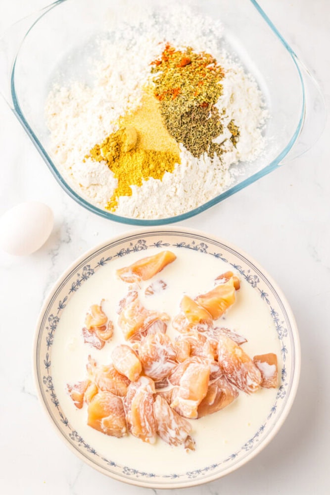 A bowl of flour and spices next to a plate of raw chicken pieces marinating in a creamy liquid, with a whole egg nearby on a white surface.