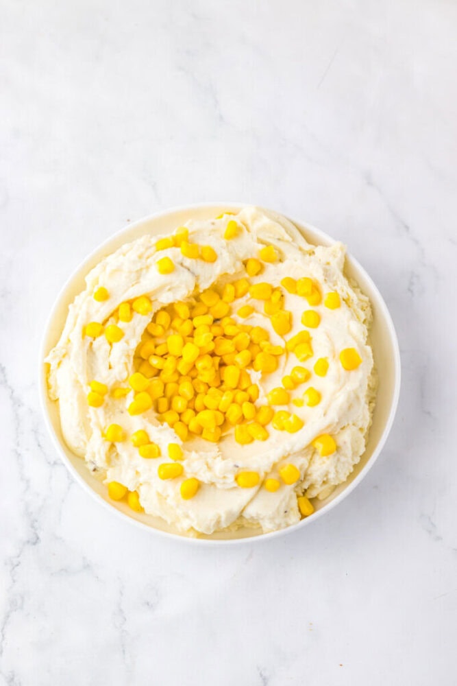 Bowl of mashed potatoes topped with yellow corn kernels on a white marble surface.