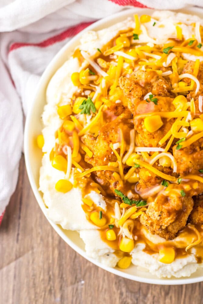 A plate of mashed potatoes topped with fried chicken pieces, shredded cheese, corn, brown gravy, and chopped herbs.